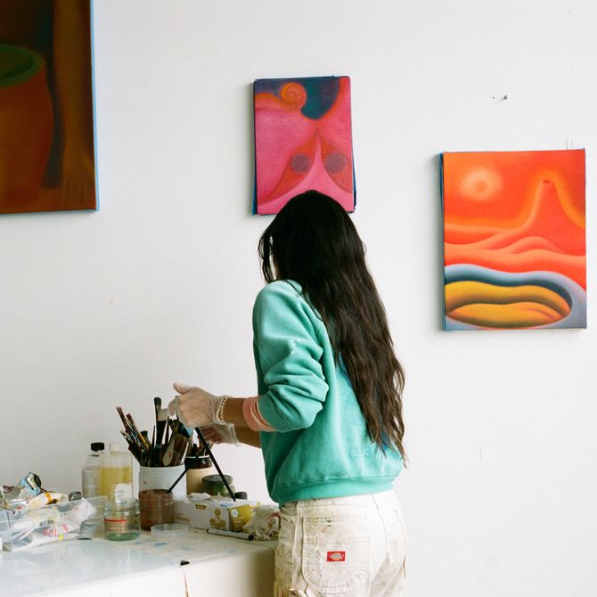 Camilla Engström in her studio getting paint supplies from a desk, three small paintings hang on the wall in the background