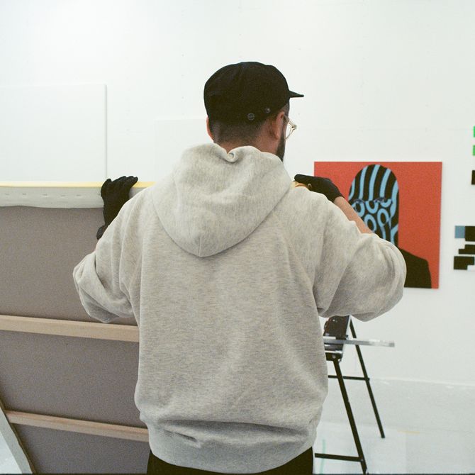 artist Shigeki Matsuyama wearing a grey hoodie and black cap, holding up a canvas in his studio
