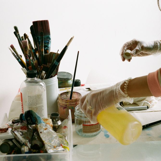 Camilla Engström pouring a liquid into a jar, preparing for painting, next to a pot of paintbrushes and a tray of paint tubes