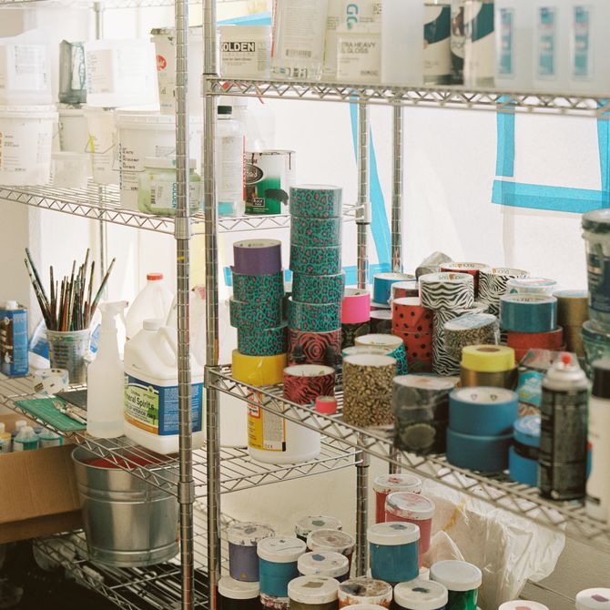 shelves in the artist's studio which are laden with stacks of different coloured and patterned tape rolls, paint pots of various colours, paint brushes, and other bottles and materials.