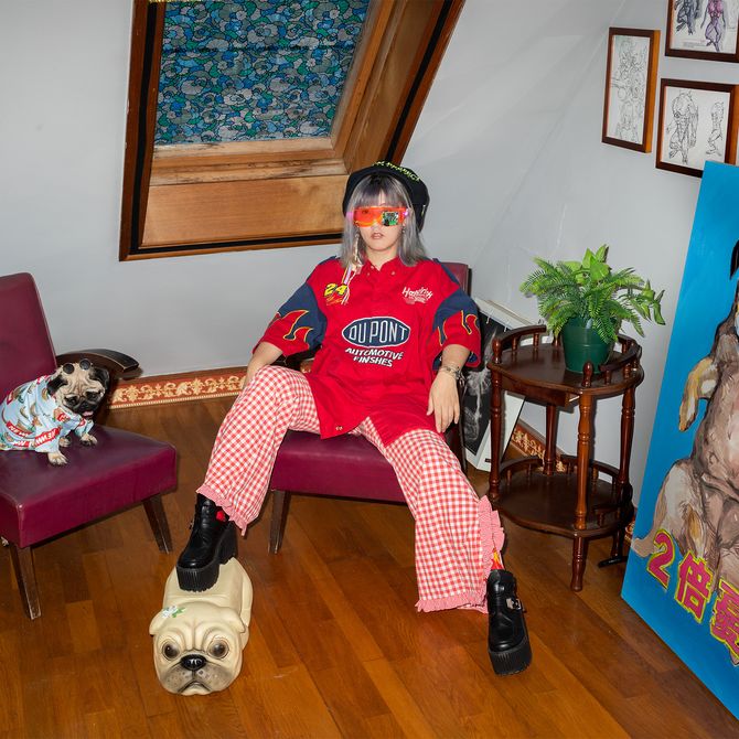 artist sitting with one leg propped on a pug stool, with their real pug on a chair next to them and a pug painting to the side