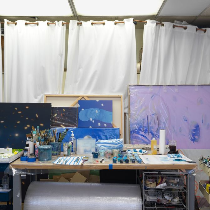 paintings on canvases stacked in front of one another behind a table of artistic tools and materials 
