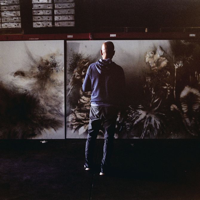 artist with his back turned to camera in a dark room gazing towards the large painting that stands before him