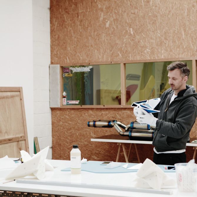 Frederic Plateus in his studio holding up an artwork whilst standing behind a large table with materials on
