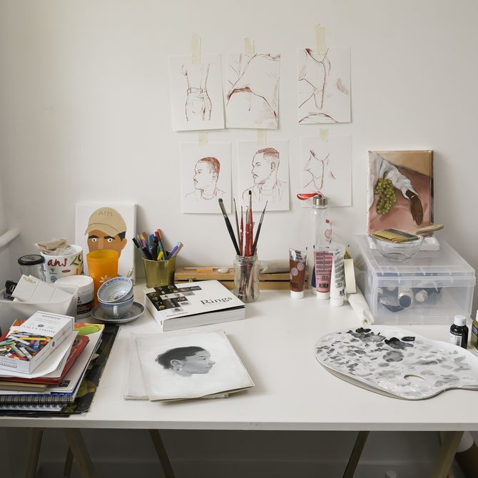 artist desk in a corner of his studio with sketches stuck up on the wall behind it, a palette with tones of black and white paint on it, notebooks and a pot of paintbrushes on it