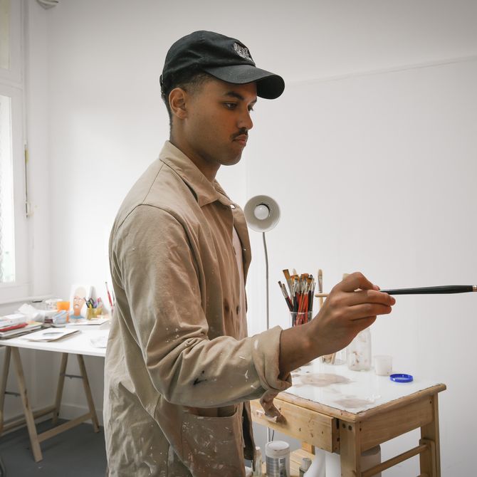 artist concentrates as he holds a paintbrush up to a painting on an easel in front of him in his studio