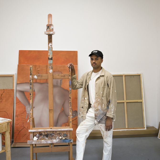 artist leaning against a wooden easel in his studio with two in-progress paintings behind him