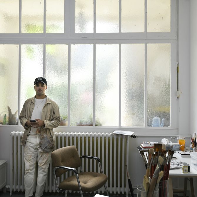 artist leaning against a radiator below a window in his studio, to the side of his desk and a desk chair
