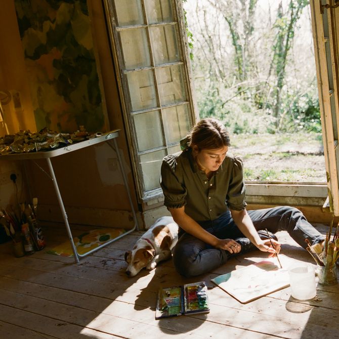 artist sat on floor with dog beside her whilst she works on a small painting
