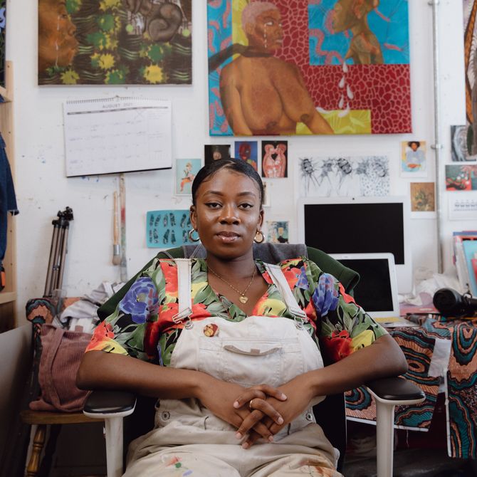 Shannon Bono sat on a chair in her studio smiling, with paintings and images taped to the wall behind her