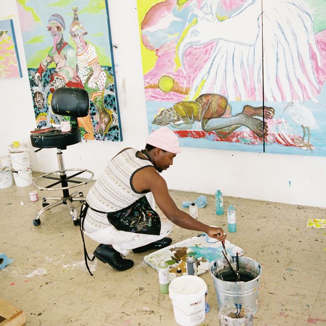 artist kneeling down to the floor to dip his paintbrush into a paint palette, with a large surrealist painting in front of him