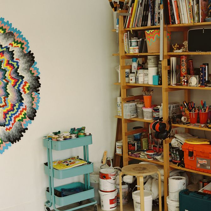 corner of the artist's studio with half of a kaleidoscopic colourful painting visible, next to a tray of paint pots, and a large unit of shelves containing paint brushes, books, pots and other materials