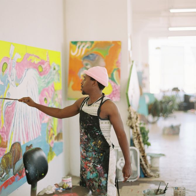 artist wearing apron covered in paint holding a paintbrush and reaching his hand towards a canvas in front of him