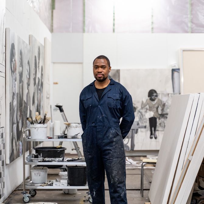 artist wears dark blue overalls and stands with hands behind his back in his studio, with empty canvases to his left and a selection of his artwork on the wall to his right