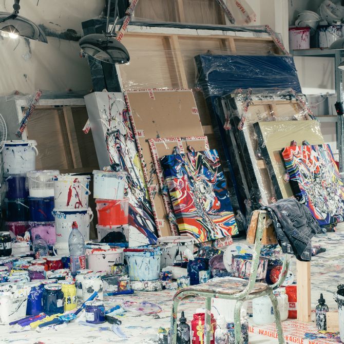 messy artist's studio with white paint covering the floor, with various paint pots on top, a chair and paintings on various sizes of canvas stacked up against one another