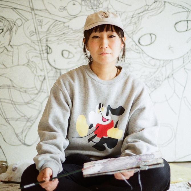 Aya Takano sitting cross-legged on the floor with a paintbrush in her hand