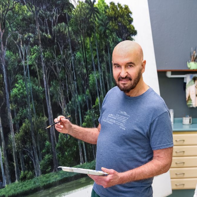 Tomas Sanchez smiling and holding a paintbrush and paint palette next to a large painting of a forest