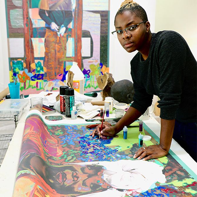Amani Lewis stood over a large painting which they hold a paintbrush to as they look to the camera