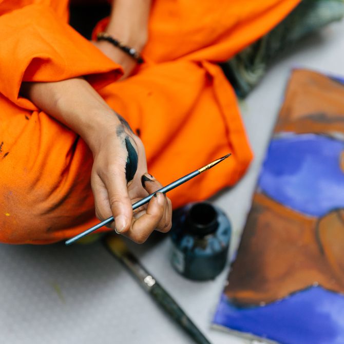 Artist sitting cross-legged on the floor holding a paintbrush dipped in black paint with a painting in front of her 