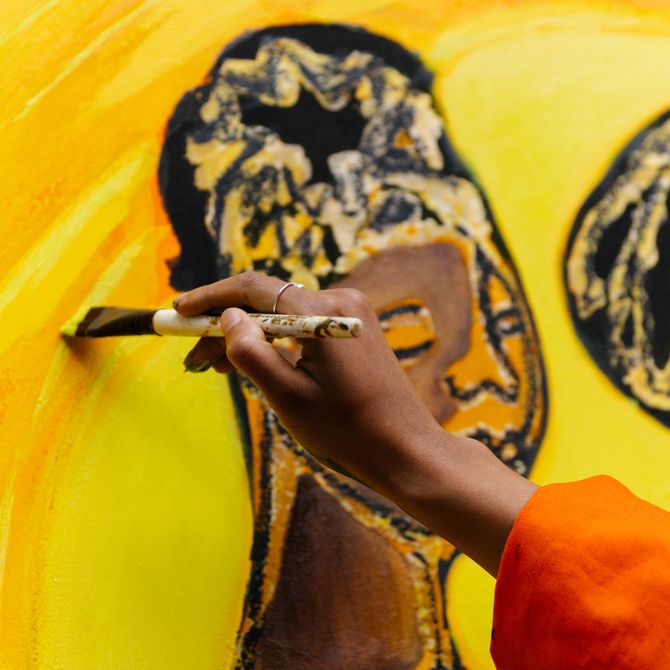 Artist holding paintbrush up to add details to a yellow painting with two figures on it