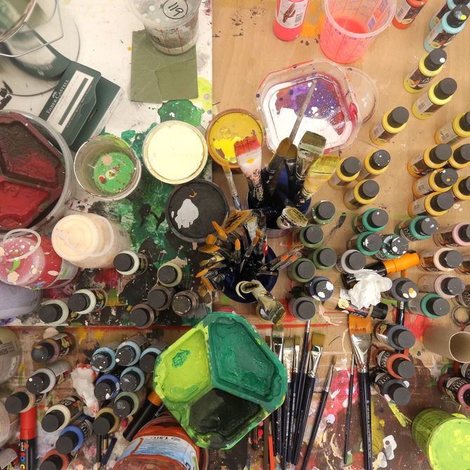a bird's eye view of an array of colourful paint pots, tubes and brushes on a wooden surface