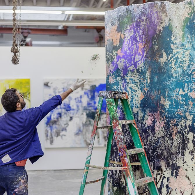 artist standing behind a ladder with arm outstretched towards large abstract painting with splashes of coloured paint on it