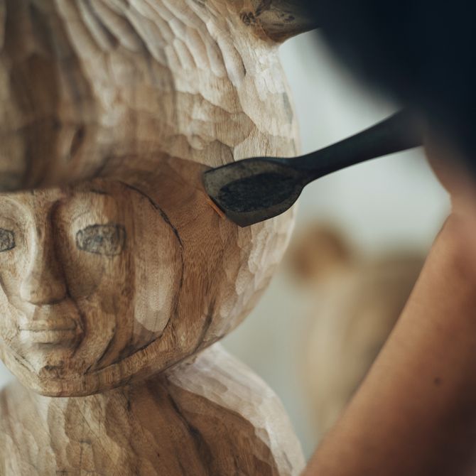 close-up of the head of a wooden sculpture with the artist working into it with a carving tool 