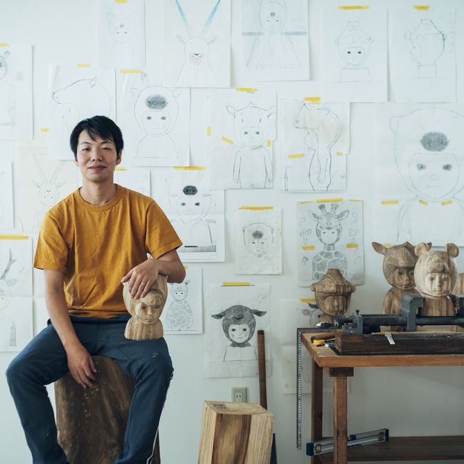 Satoru Koizumi sat on a block of wood with his arm resting on a small wooden sculpture placed on his knee, in front of a wall filled with pencil sketches on paper