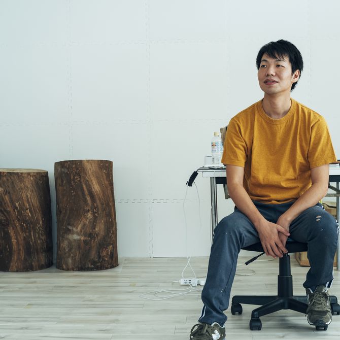 Satoru Koizumi wearing an orange t-shirt sat on a chair in his studio
