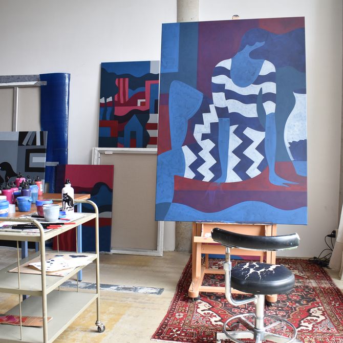 Parra's studio, without him. A large painting takes centre stage, showing a faceless blue woman in a striped dress, painted in red, purple, blue and teal. The studio is full of brightly coloured paints, with a large window on the right and a patterned rug across the floor under the painting. 