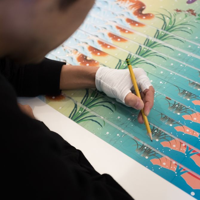 artist hand holding yellow pencil signing bottom of multiple works of art layered on table