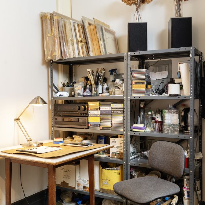 artist studio with bright lamp on desk with desk hair and shelves of paint brushes, books, videos, paintings and two vases of flowers