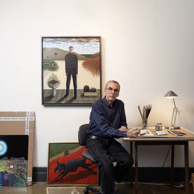 Scott Kahn at his desk with a paintbrush in hand and three of his paintings in the background