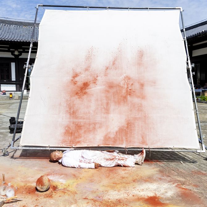 Artist lying down on ground in front of white stretched canvas with red marks on it outdoors