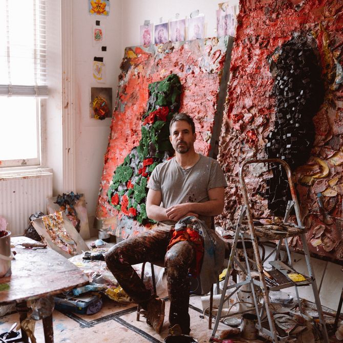Antony sitting in his studio in front of two large textured paintings leaning against the wall