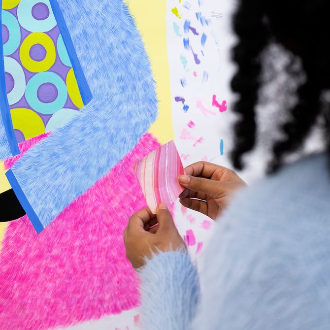 artist holding up a small square of pink paper in front of a colourful painting