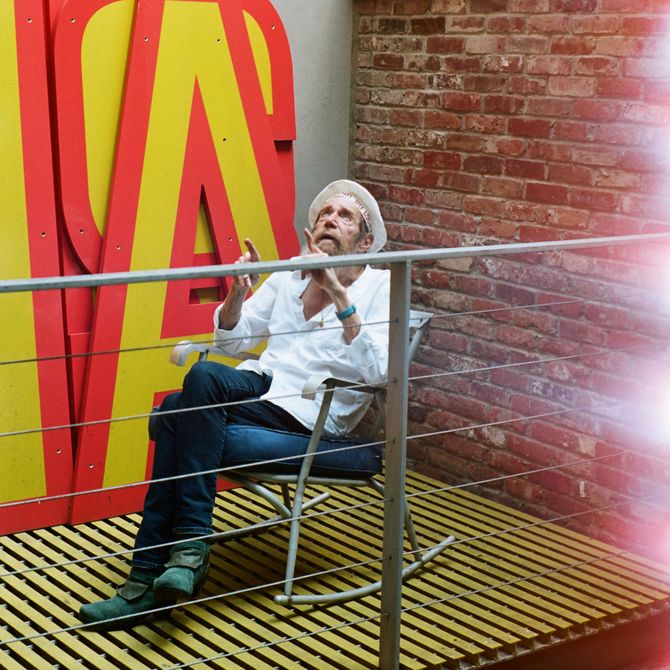 Lawrence Weiner sat outside on a metal rocking chair in front of some large yellow and red letters. He is looking at the sky.