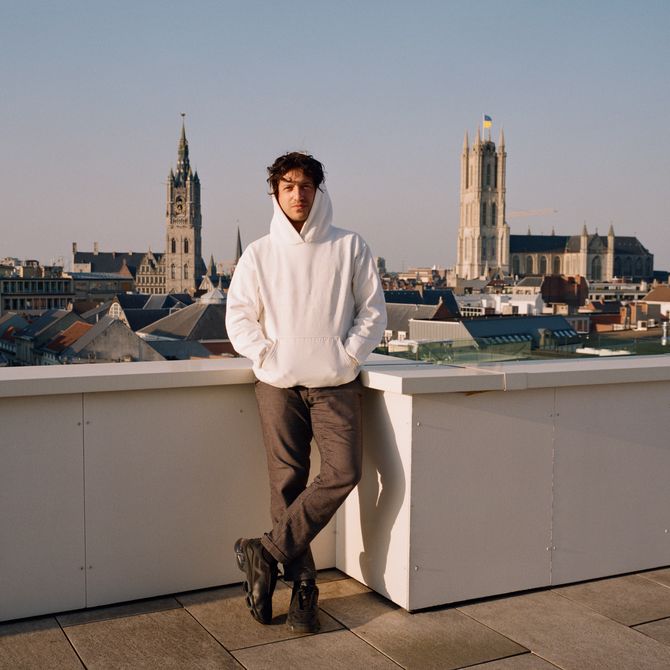 David Rudnick wearing a white hoodie on the roof of his studio, a panoramic view of Ghent's skyline is visible behind him