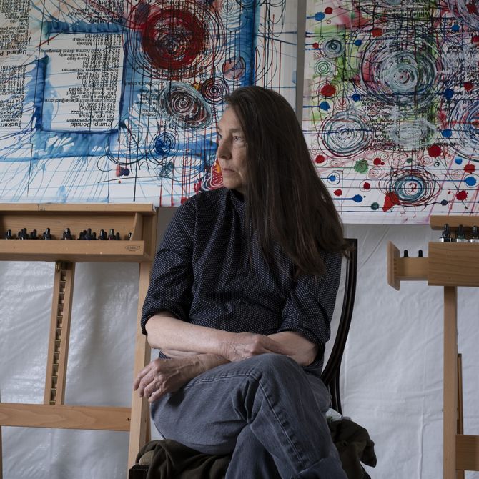 Jenny Holzer sitting in her studio, glancing to the side, with two paintings on easels behind her