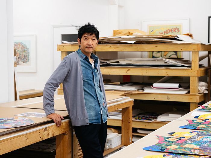 artist in paint-splattered denim shirt, leaning on a table on which a row of colourful prints are laid