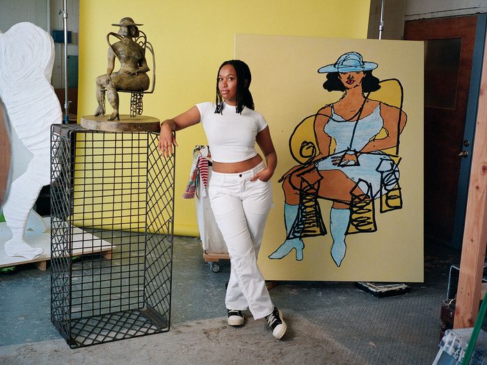Tschabalala Self in her New Haven studio, wearing white and posing with a bronze sculpture and large yellow painting