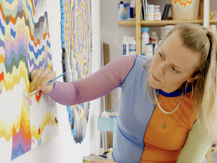 artist wearing colourful long-sleeved top concentrating on an unfinished painting in front of her which she holds a small paintbrush to