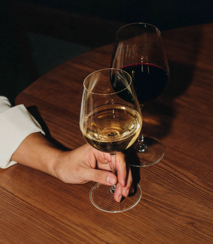 Hand with a glass of white wine at a table, with a glass of red wine in the background.