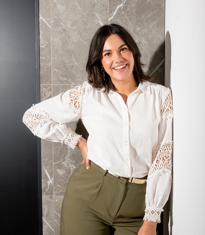 Vrouw met donker haar glimlacht in witte blouse met kant en groene broek, leunend tegen een marmeren muur.