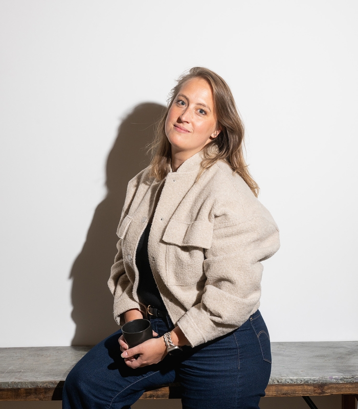 Vrouw zittend op een bank met een mok in haar hand, draagt een beige jas en blauwe broek, tegen een witte achtergrond.