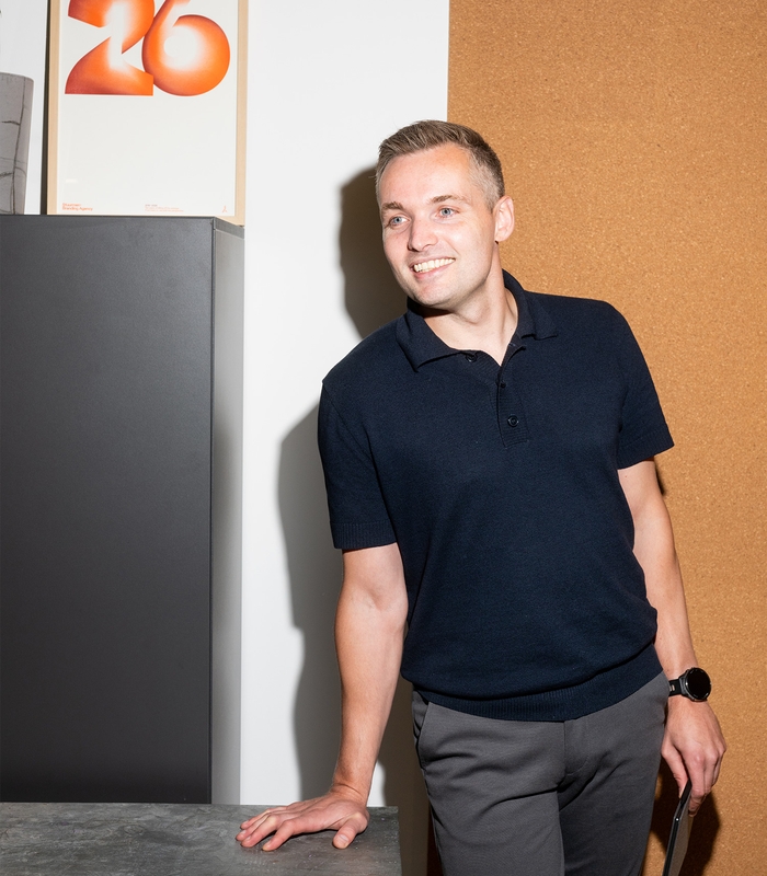 Man in donkerblauwe polo en grijze broek leunt tegen een bureau. Achtergrond met poster van het nummer 26 en kurkwand. Lachend gezicht.