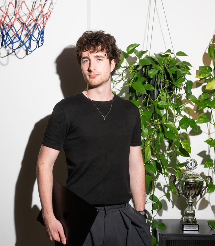 Man met krullend haar houdt een laptop vast in een kamer met planten, een bokaal en een basketbalnet op de achtergrond.