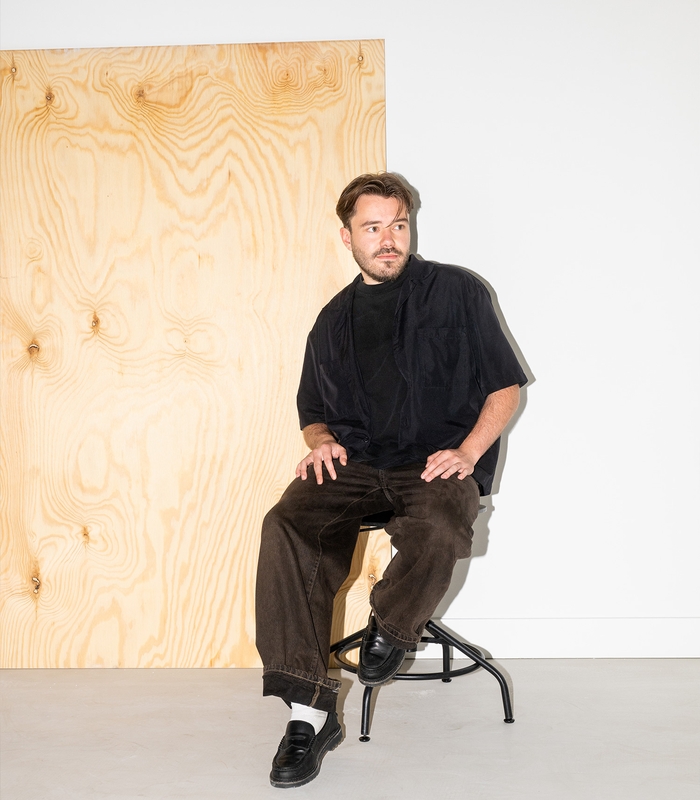 Man zit op een kruk in een minimalistisch ingerichte ruimte met een houten paneel en witte muren, gekleed in zwart shirt en bruine broek.