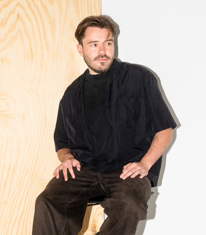 Barend met lichte baard zit voor een houten paneel, draagt een zwart overhemd en kijkt naar links. Achtergrond is wit en houtkleurig.