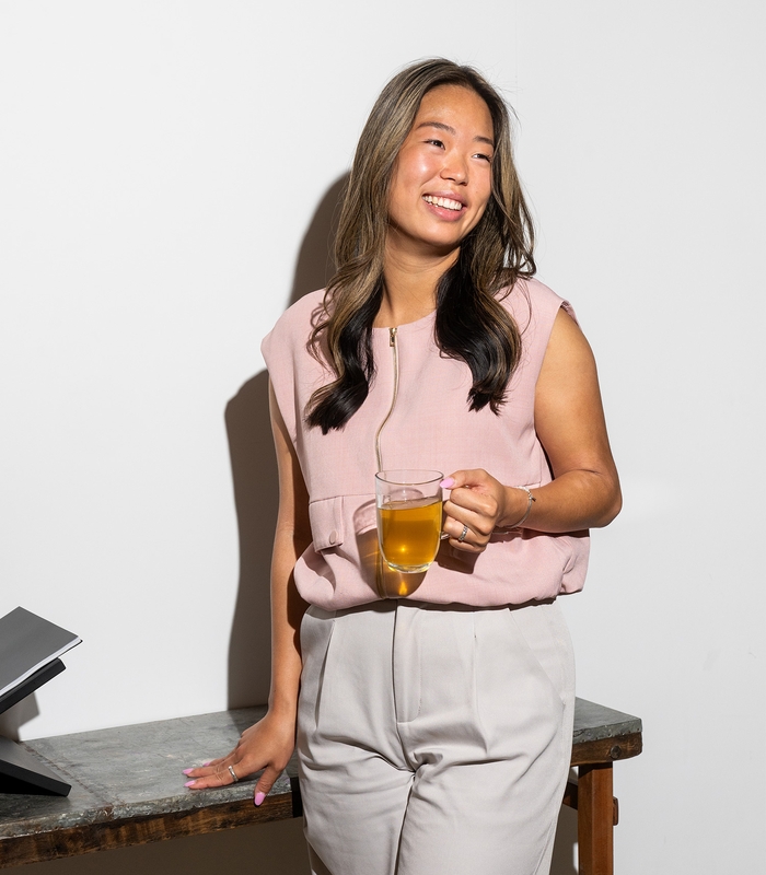 Maria in een roze blouse en grijze broek leunt lachend tegen een tafel, met een glas thee in haar hand.